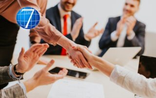 A business meeting scene showing a confident handshake between two professionals, with colleagues in the background clapping and smiling in support, symbolizing the concept of leading by example and fostering a positive organizational culture.