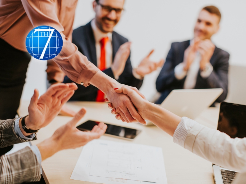 A business meeting scene showing a confident handshake between two professionals, with colleagues in the background clapping and smiling in support, symbolizing the concept of leading by example and fostering a positive organizational culture.