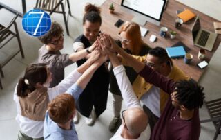 A diverse group of professionals standing in a circle and joining hands in a celebratory high-five, symbolizing teamwork, small wins, and success. The background features a modern office setup with a wooden table, laptops, and office supplies, highlighting a collaborative and productive work environment.