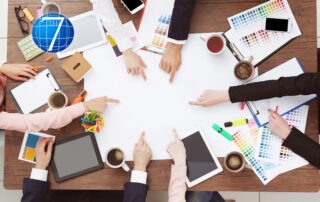 A collaborative team working around a table, pointing at a blank space in the center, surrounded by tools like color swatches, tablets, notebooks, and coffee cups. This image symbolizes brainstorming and innovation, representing the essence of a learning organization.